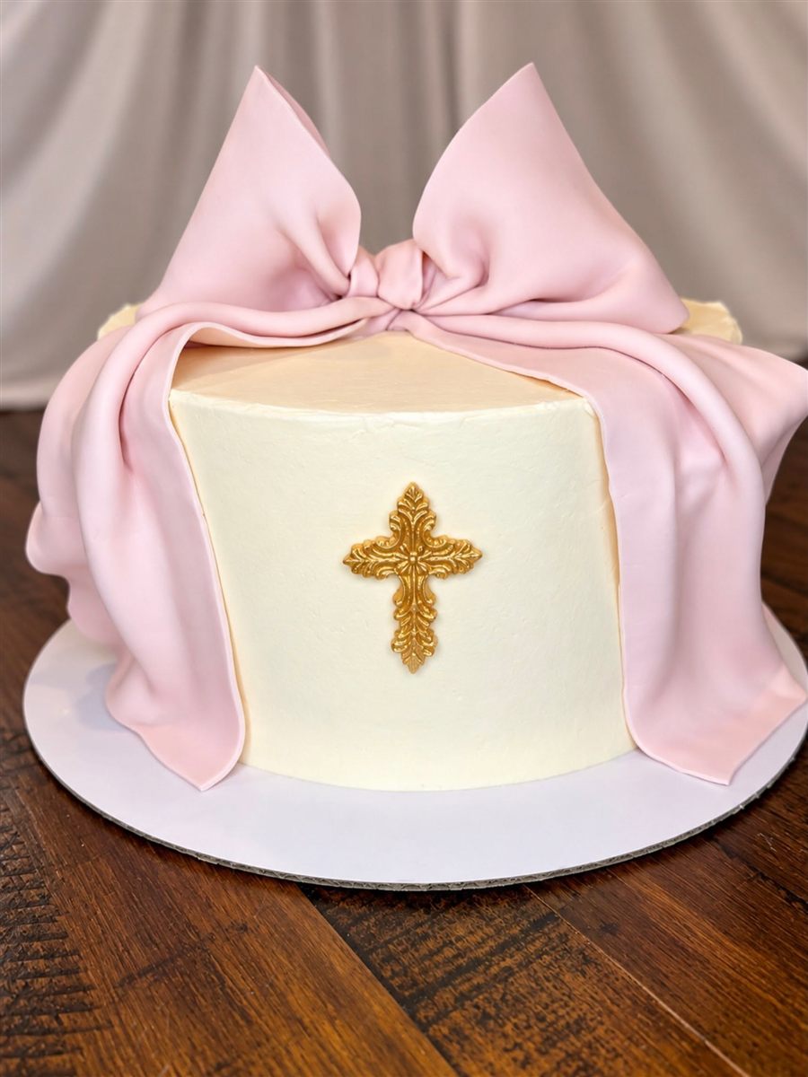 White cake wrapped in a pink fondant bow with a gold cross
